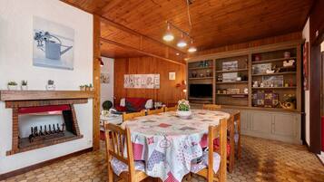 A cozy dining area with a wooden table, chairs, a brick fireplace, and a kitchen with shelves.