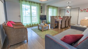 A living room with a sofa, two wicker chairs, a TV, a dining table with chairs, and a kitchen area. - Apartment 'Le Bois De Marie' with Mountain View, Private Terrace and Wi-Fi (Barèges)