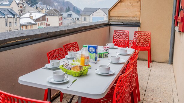 A balcony with a table set for breakfast, including cups, bowls, and a basket of food. - Apartment 'Le Bois De Marie' with Mountain View, Private Terrace and Wi-Fi (Barèges)