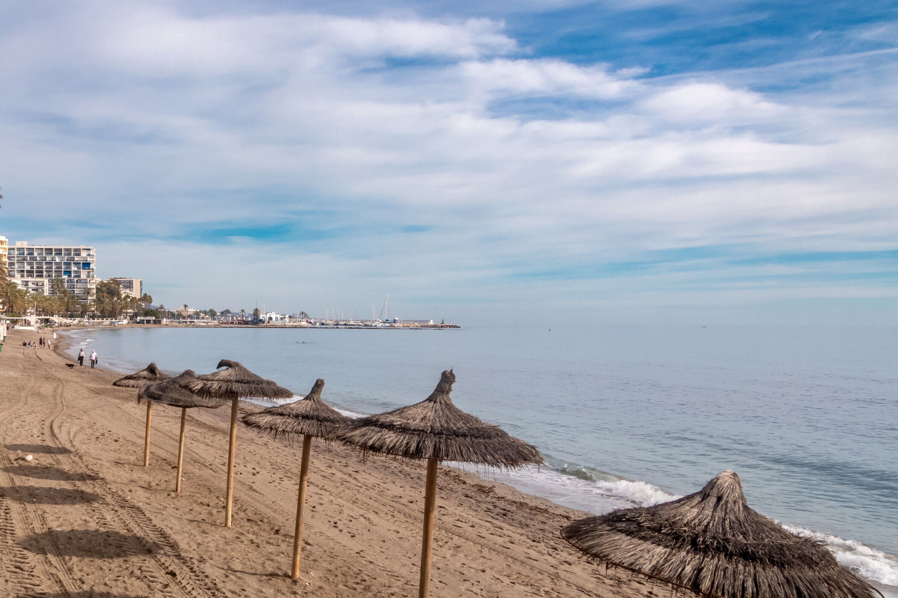 A beach with thatched umbrellas, a sandy shore, and buildings in the background.