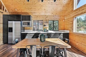 Interior de una cabaña de madera con cocina, mesa de comedor, sillas y un ventanal grande.
