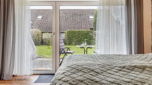 Un dormitorio con cama, puerta corrediza de vidrio que da a un patio con mesa y sillas, y vista a una casa con techo de tejas.