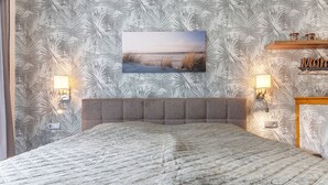 Un dormitorio con cama, cabecera, lámparas fijadas a la pared, un cuadro y una estantería con adornos.