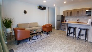 A compact living space with a sofa, two armchairs, a small coffee table, a potted plant, a kitchen area with a refrigerator, and a microwave.