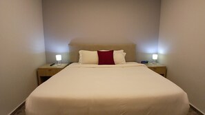 A neatly made bed with white bedding and a red accent pillow, flanked by two bedside tables with lamps and a digital clock.