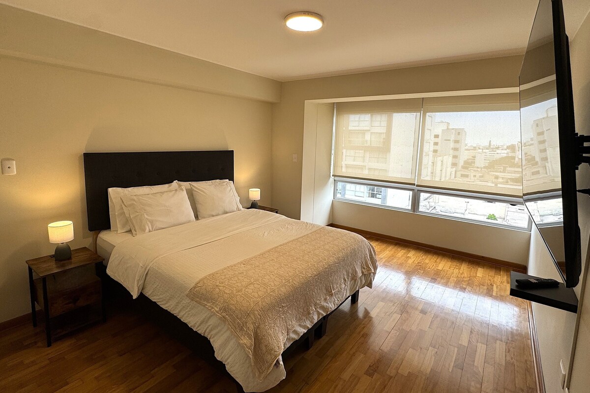 Une chambre à coucher avec un lit, des lampes de chevet, une fenêtre donnant sur des immeubles et un téléviseur posé sur un meuble.