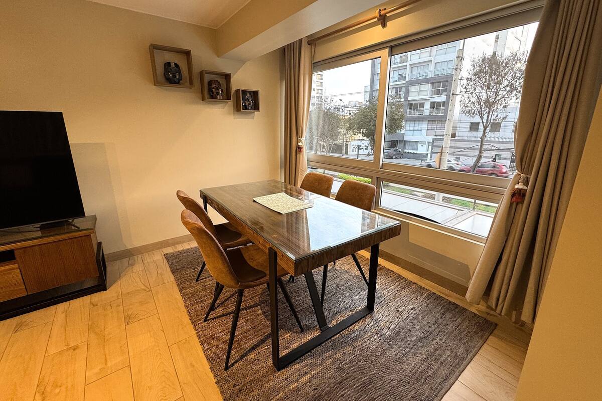 Una sala moderna con una mesa de comedor de madera, dos sillas, un televisor de pantalla plana y un gran ventanal con cortinas.