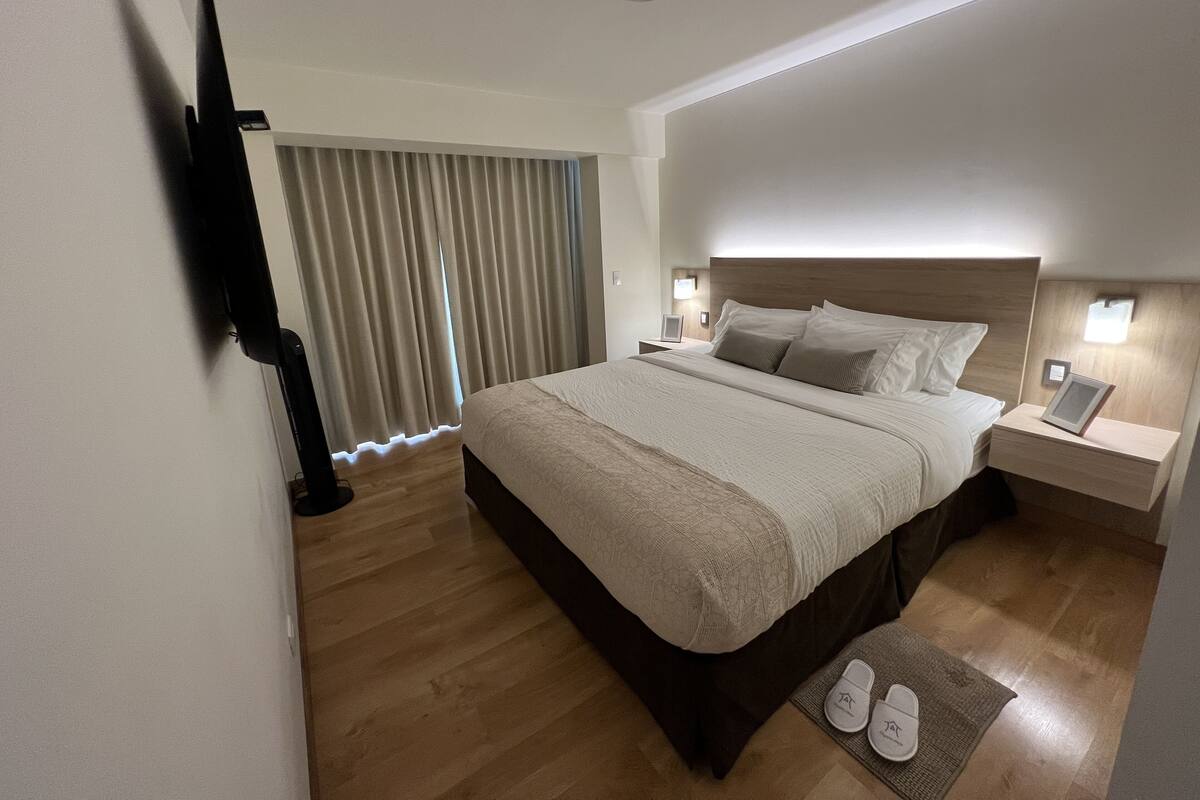 Una habitación de hotel moderna con una cama grande, mesitas de noche, un televisor de pantalla plana y vistas al exterior a través de amplios ventanales.