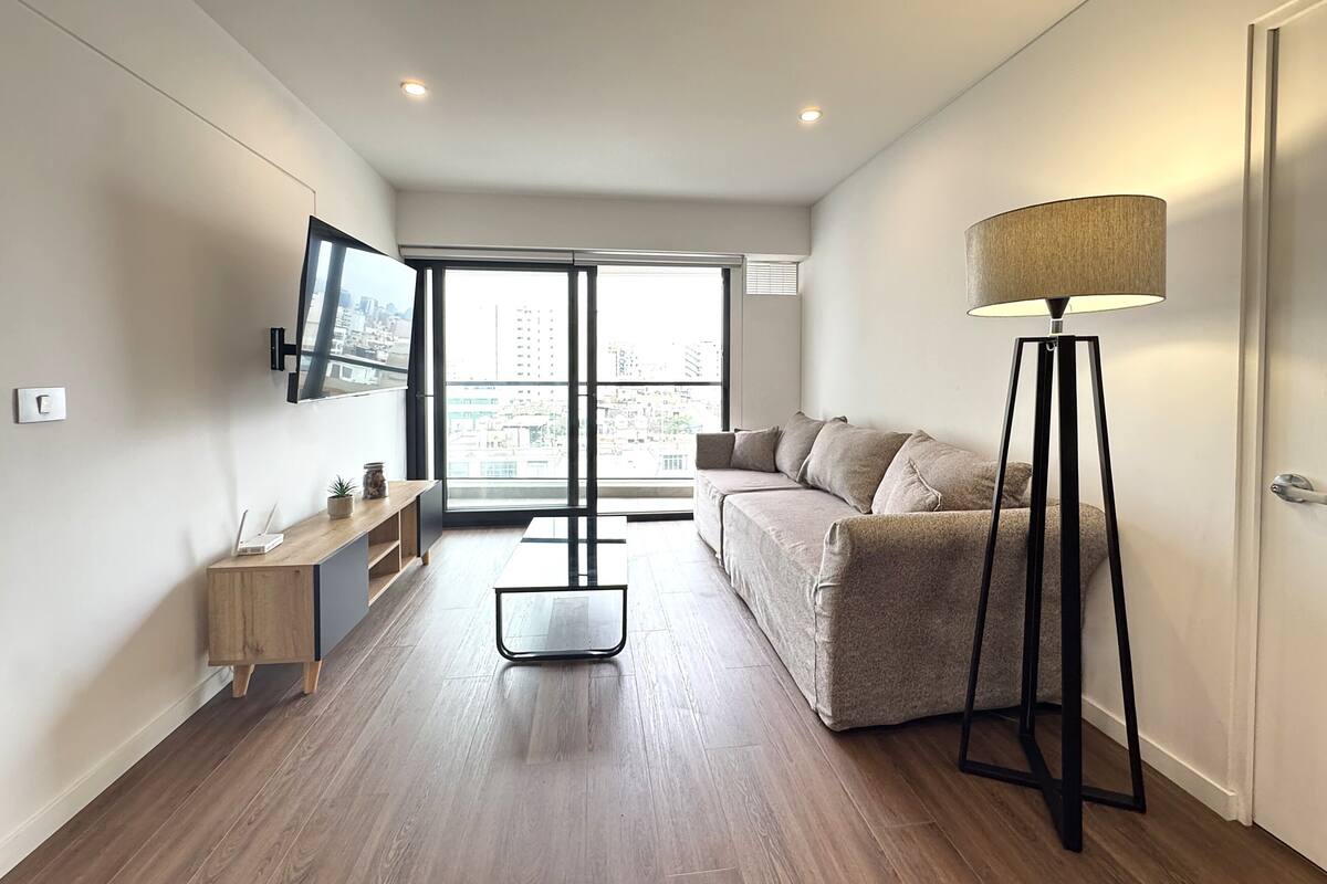 Una sala de estar moderna con un sofá, una lámpara de pie, un televisor montado en la pared y una ventana con vistas a la ciudad.