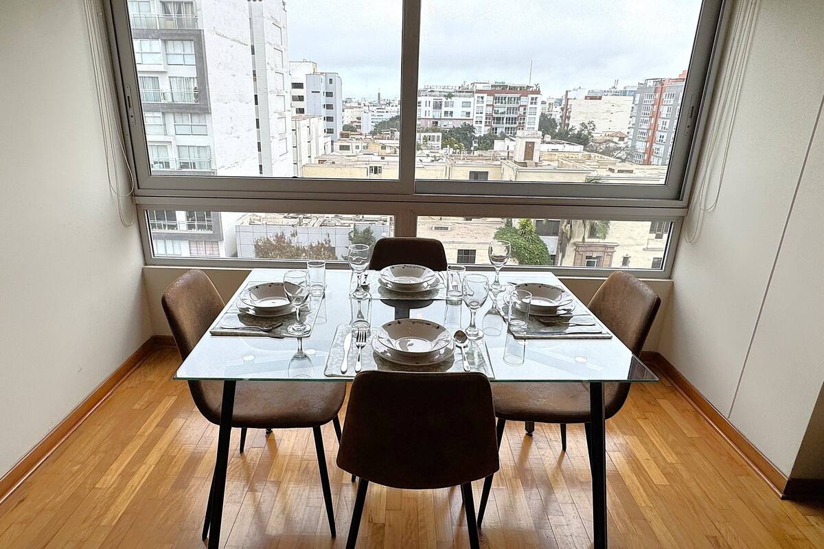 Comedor con mesa dispuesta para cuatro personas, ventanas amplias con vistas a la ciudad y suelo de madera.