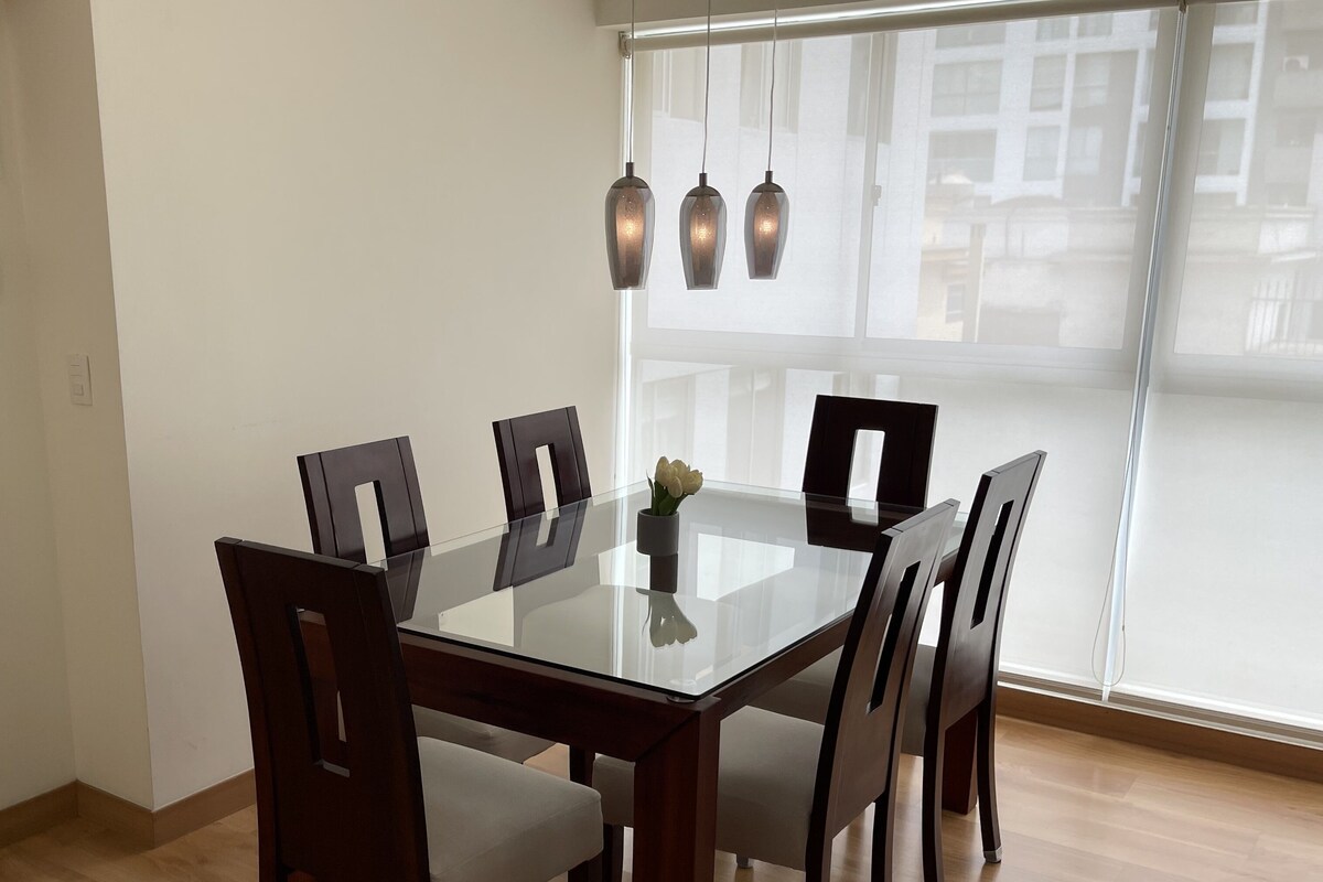 Un coin repas avec une table en verre entourée de six chaises, un vase de fleurs et des luminaires suspendus.