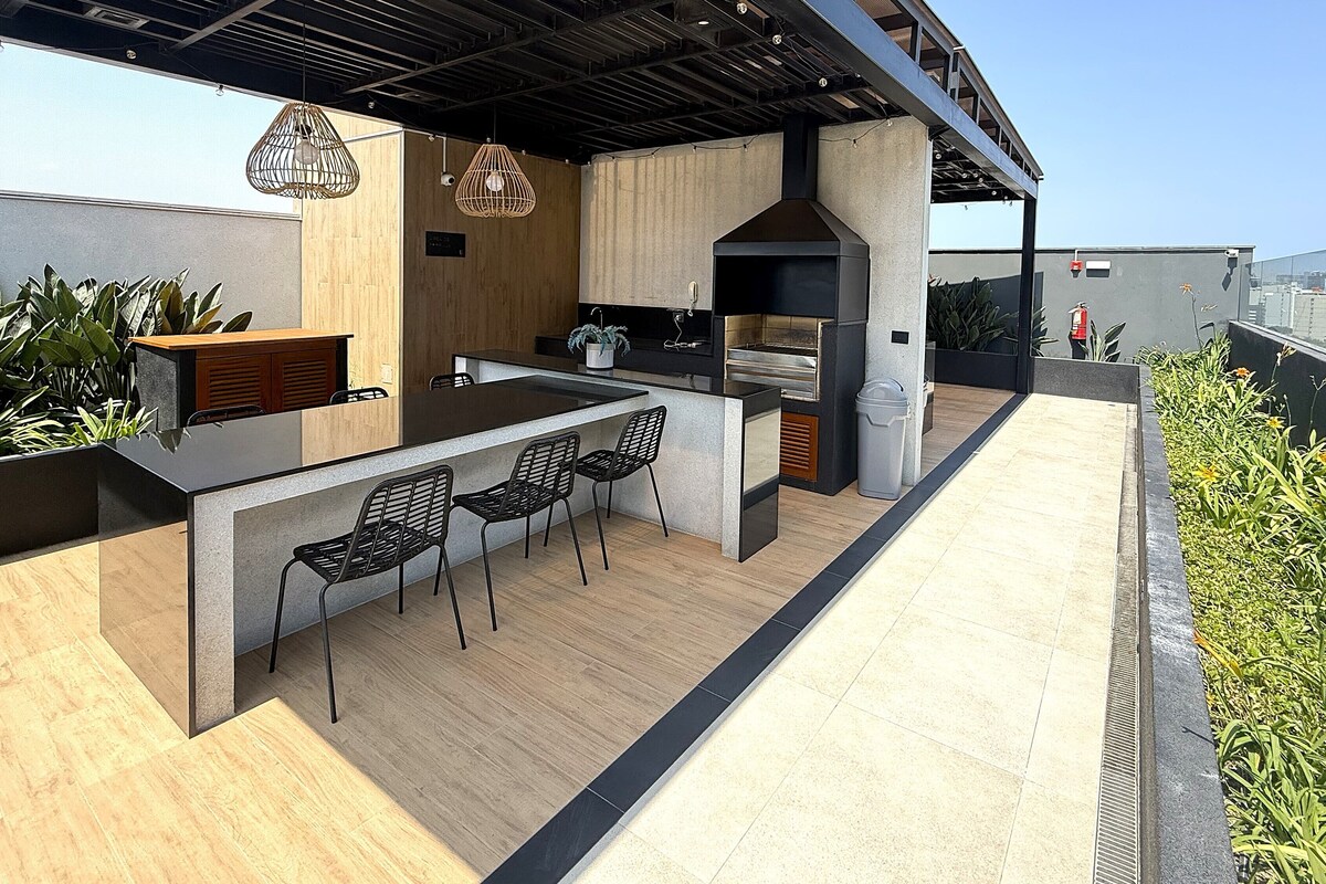 Eine Dachterrasse mit Holzboden, schwarzen Metallstühlen, einer Bar, hängenden Lampen und Blick auf die Skyline der Stadt.