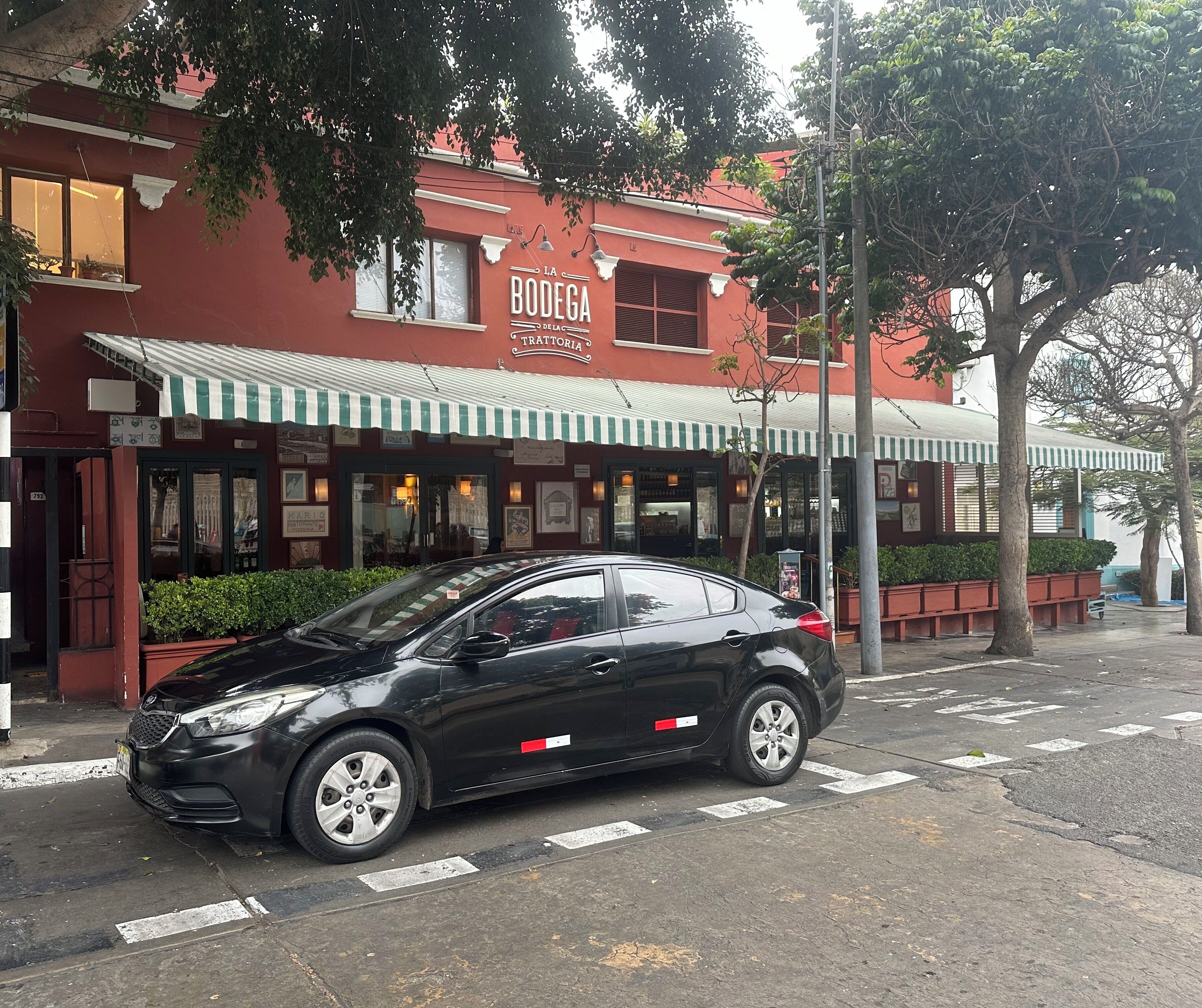 Une voiture noire est garée devant un immeuble rouge, surmonté d’un auvent à rayures vertes et blanches.