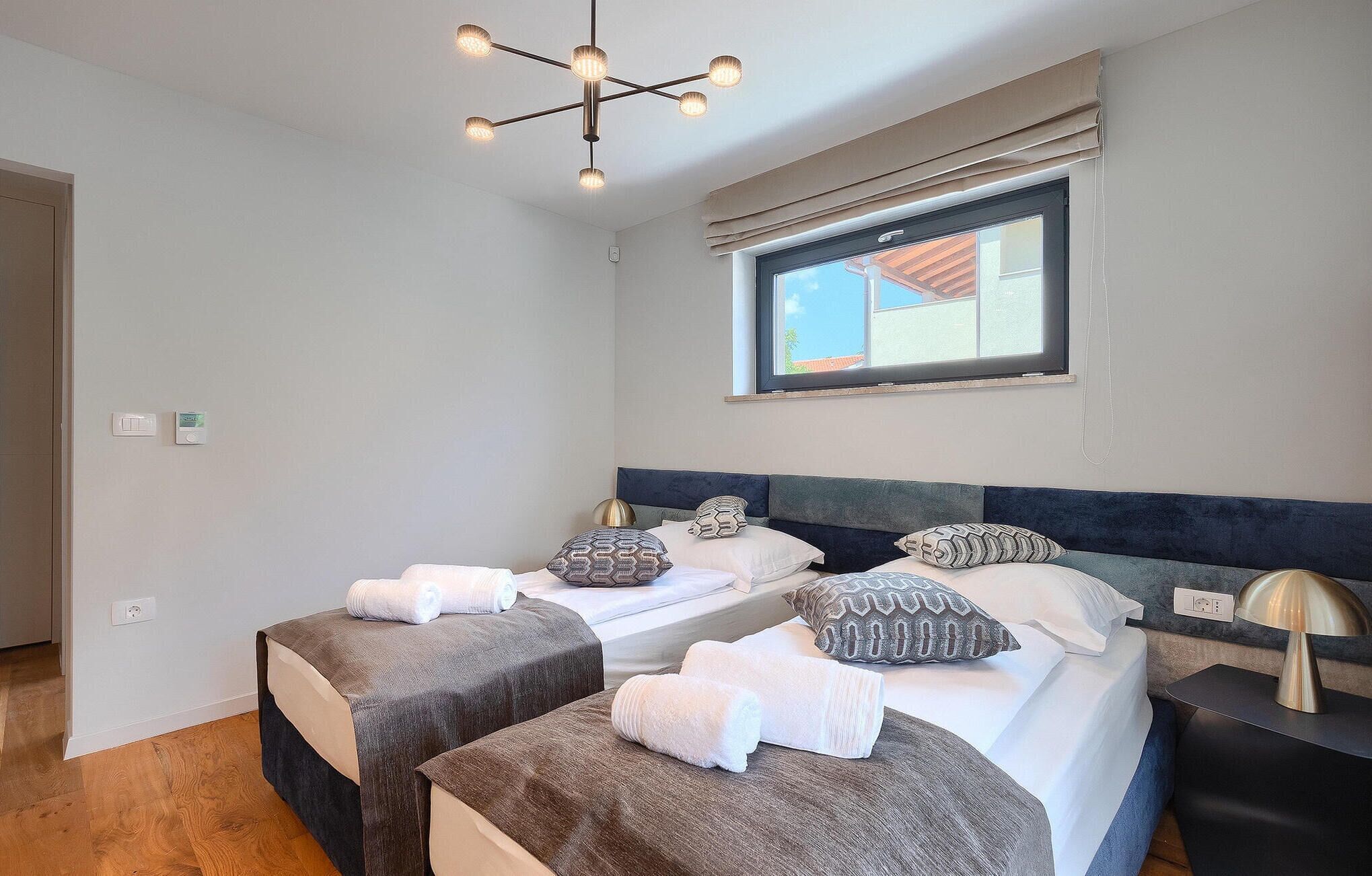 Quarto moderno com duas camas, luminária no teto, janela com vista e mesa de cabeceira com abajur.