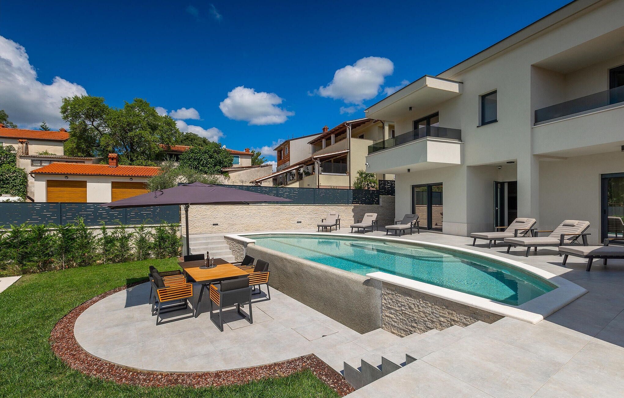 A modern outdoor pool area with lounge chairs and a table, surrounded by a well-maintained lawn and a stone wall.