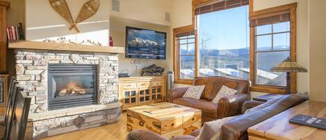 A cozy living room with a stone fireplace, leather furniture, and large windows offering a view of the mountains.