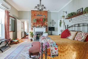 A cozy bedroom with a brick fireplace, a bed with patterned bedding, a red chair, and a colorful rug.