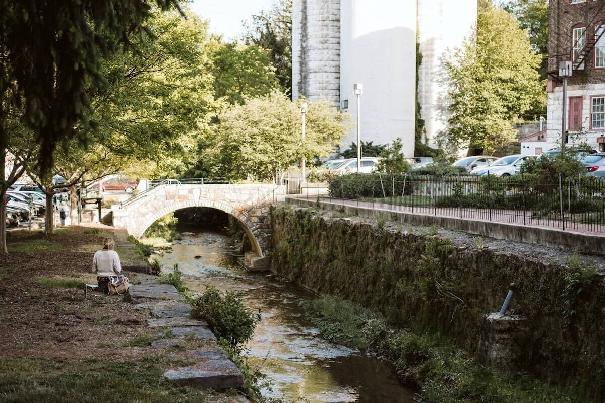 Un pont de pierre enjambe un petit ruisseau dans un parc, avec des arbres et un bâtiment en arrière-plan.