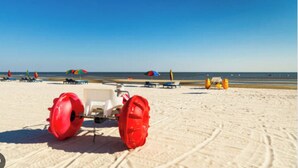 A sandy beach with a white vehicle and red wheels, umbrellas, and lounge chairs in the background.