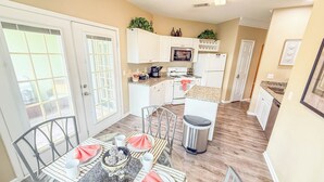 A modern kitchen with white cabinets, a microwave, stove, and a sink. There is a dining area with a table set for two, chairs, and a coffee maker.