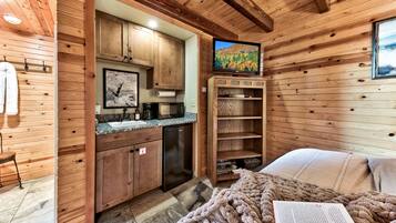 A cozy cabin-style room with wooden walls, a kitchenette, a bed with a blanket, and a television.