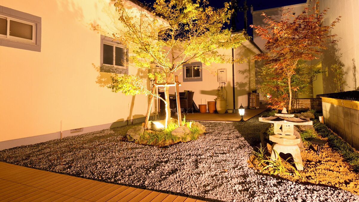 A well-lit garden area with a wooden deck, a small pond with a bridge, and a stone lantern.