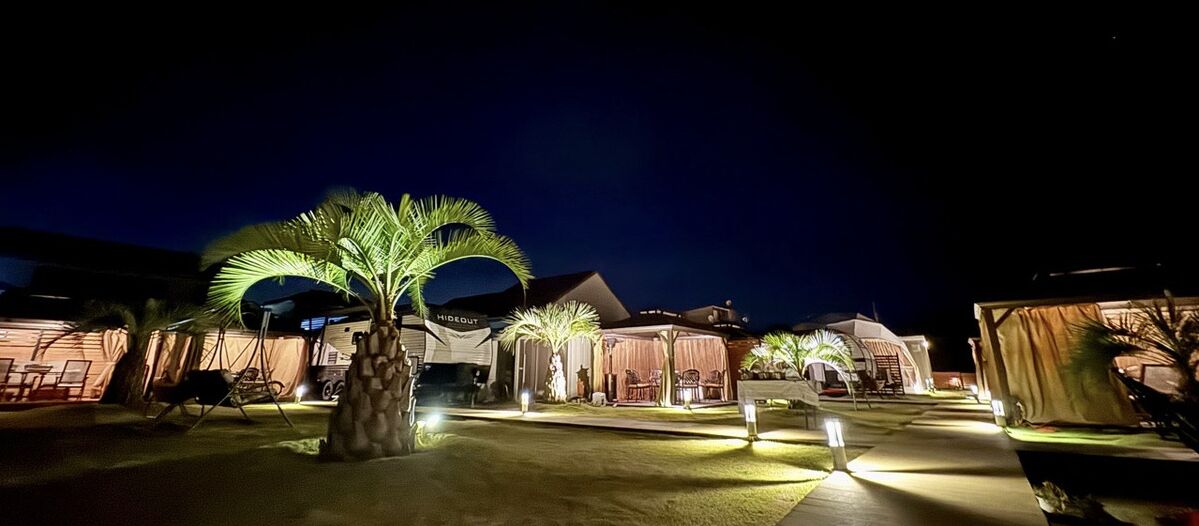 Un espace extérieur bien éclairé, agrémenté de palmiers, avec un coin salon abrité et un bâtiment sur lequel on peut lire l’inscription « RESORT ».