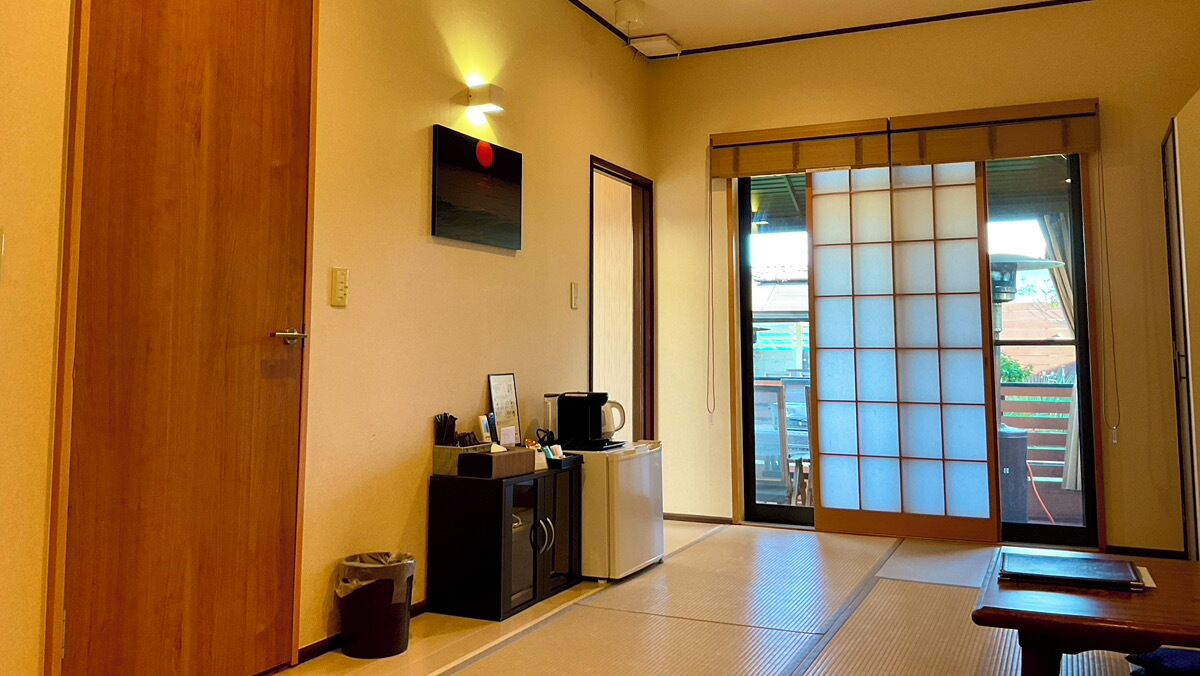 Una habitación japonesa tradicional con suelo de tatami, puertas correderas, una pequeña kitchenette y vistas a través de la puerta.
