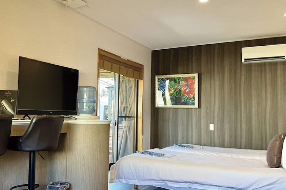 A hotel room with a bed, a desk with a computer, a chair, a large window, and an air conditioning unit.