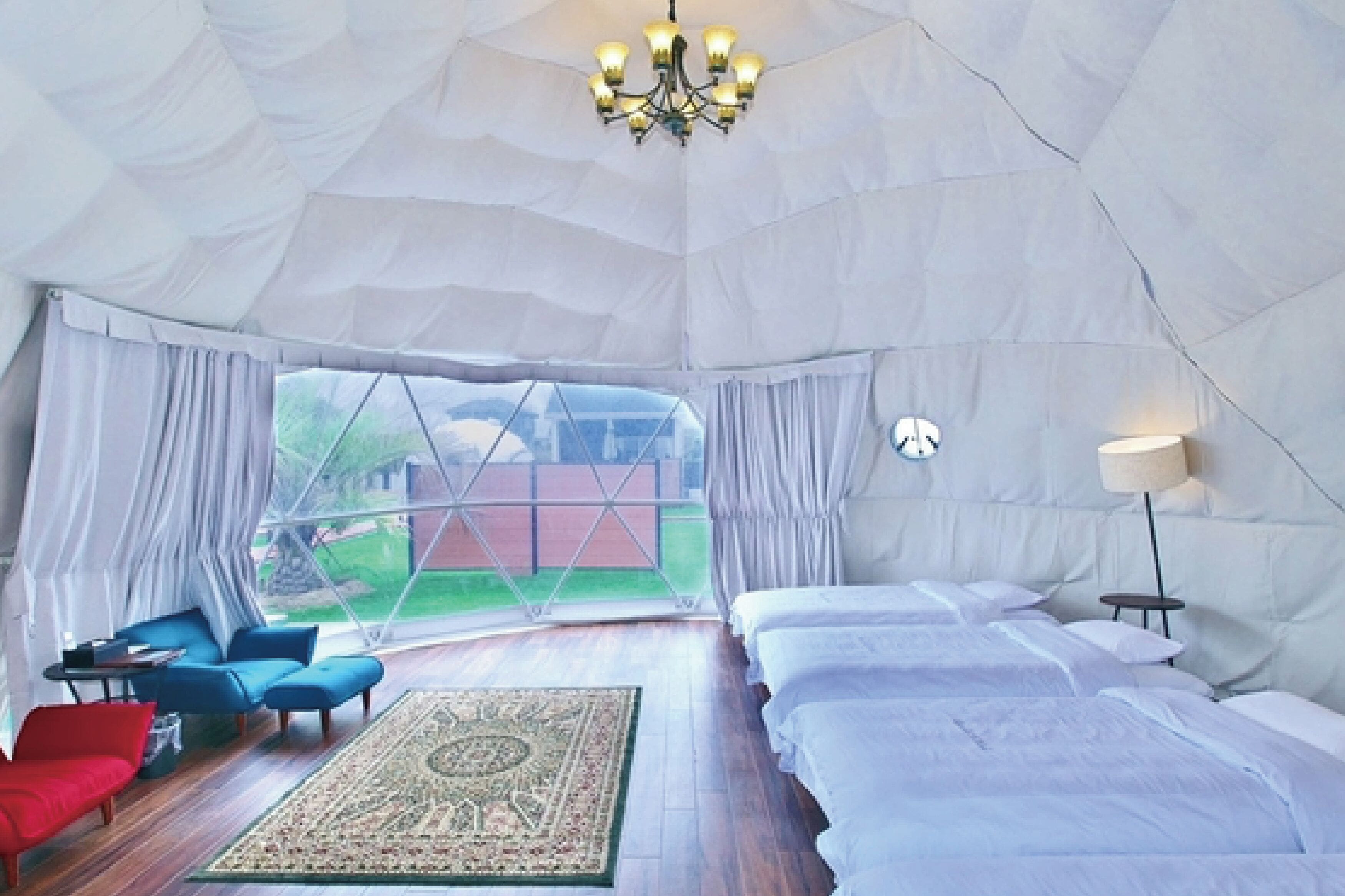 A tent-style room with a bed, two blue chairs, a red chair, a small table, a lamp, and a chandelier.