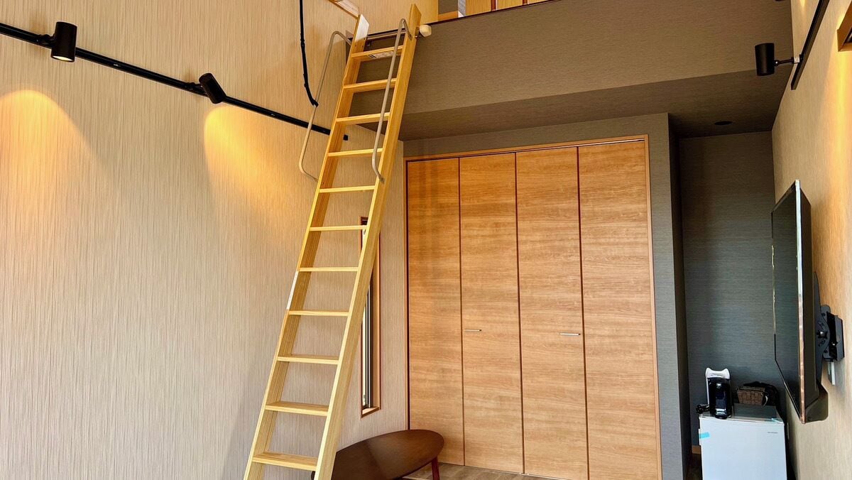 Quarto com guarda-roupa de madeira, escada, cadeira e TV de tela plana presa na parede.