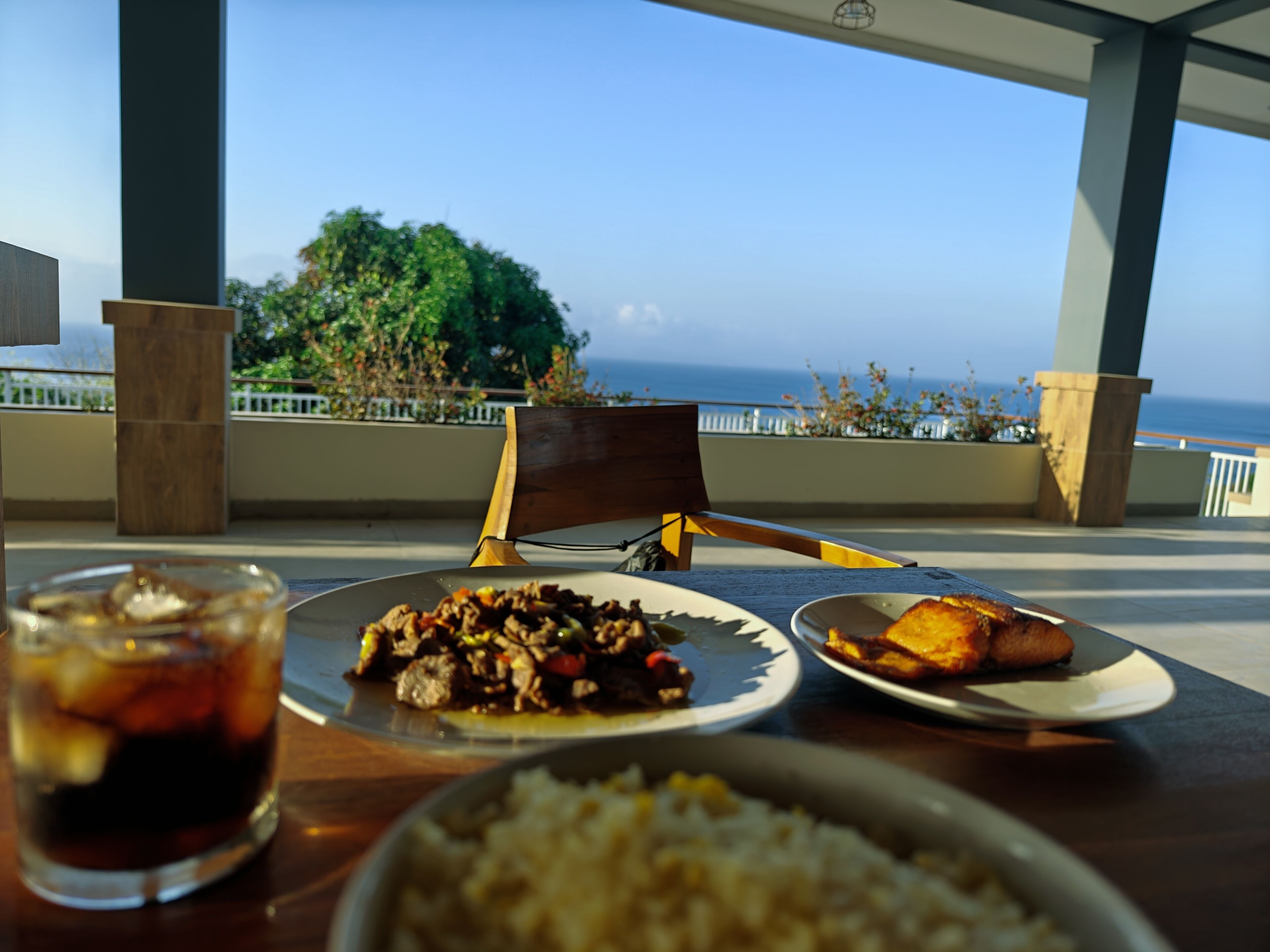 A dining table with plates of food, a glass of drink, and a view of the ocean.