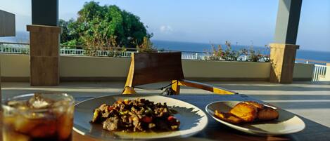 A dining table with plates of food, a glass of drink, and a view of the ocean.