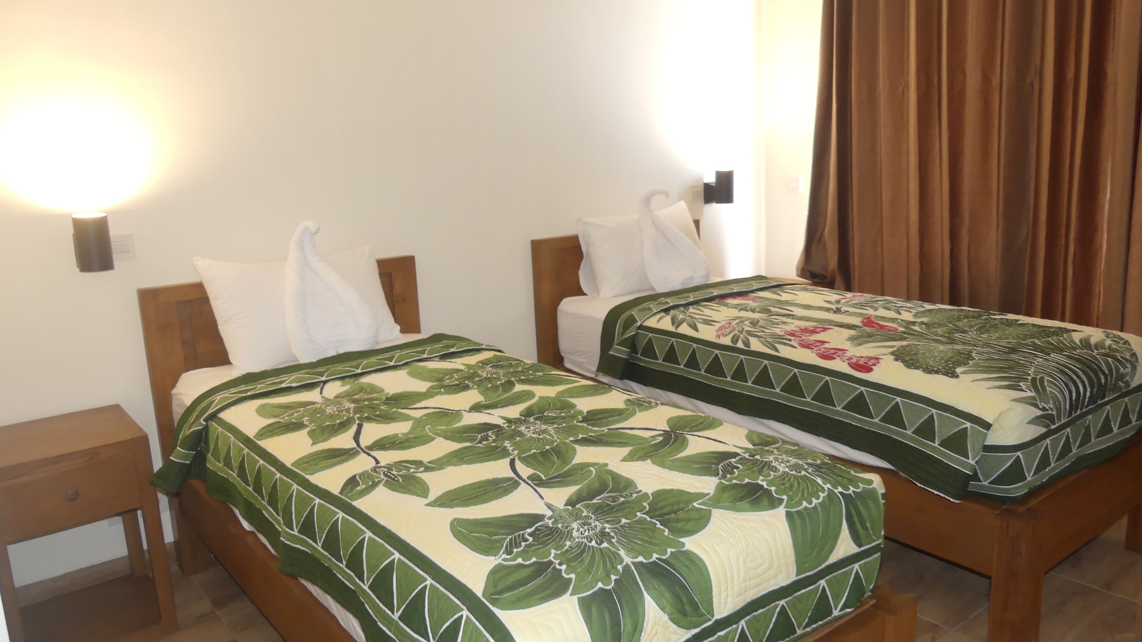 A double room with two beds, each with a floral bedspread, wooden headboards, and white pillows. There is a nightstand on each side of the beds, and a wall-mounted light fixture above the beds.