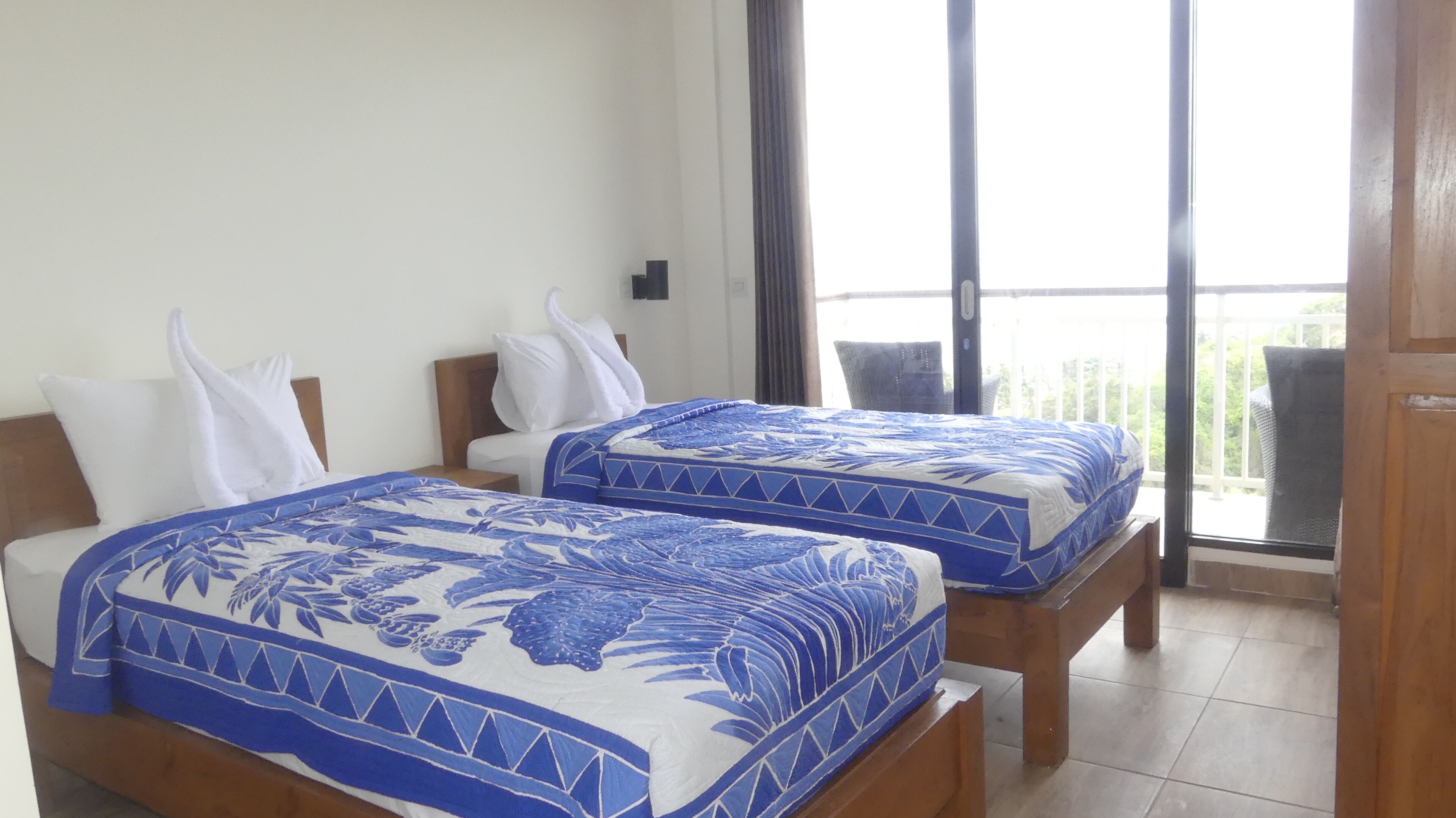 A hotel room with two beds, each with a blue and white patterned bedspread, a wooden headboard, and a view of a balcony with outdoor furniture.