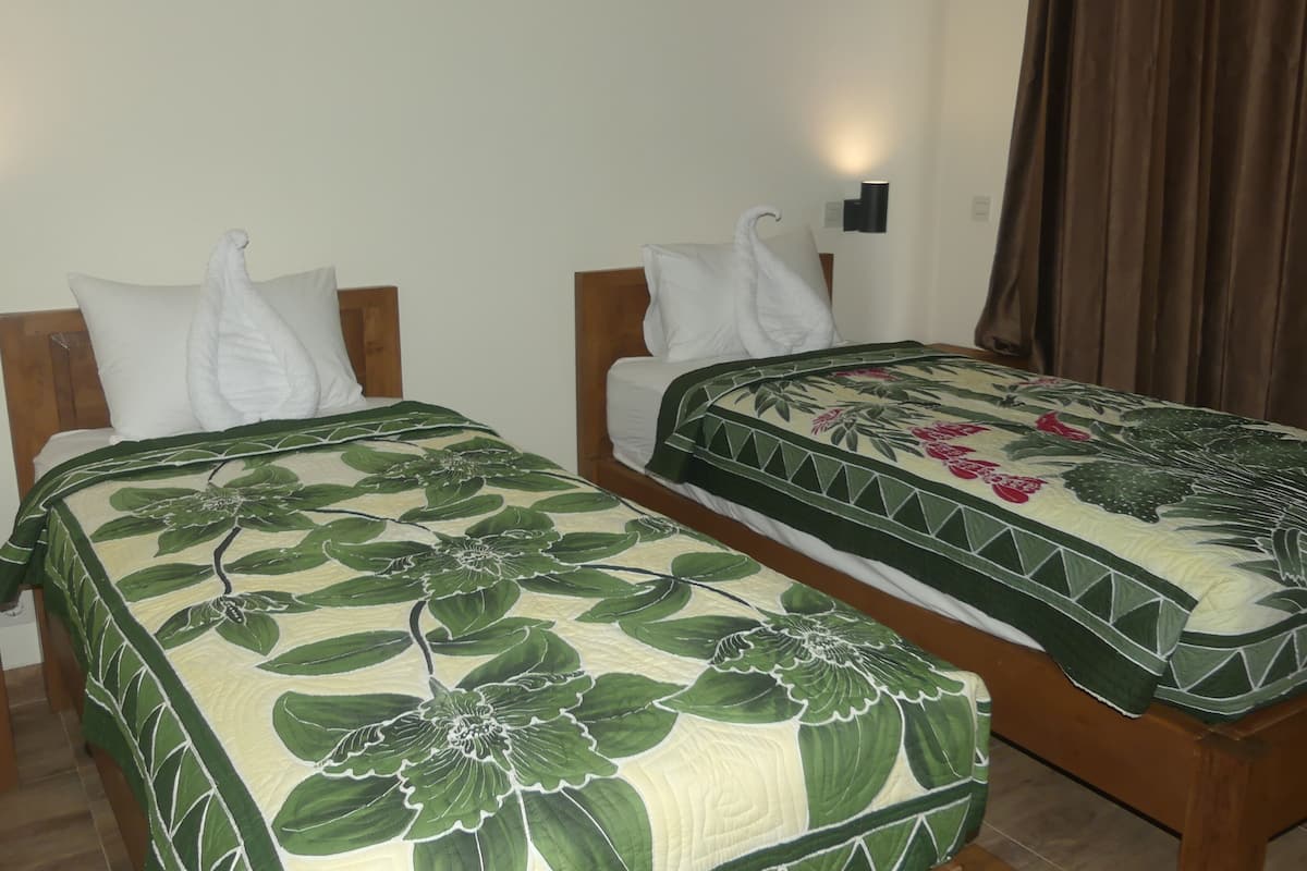 A hotel room with two single beds, each with a wooden headboard, white pillows, and a green floral bedspread. There are two wall-mounted nightstands with a wooden finish, and a brown curtain on the window.