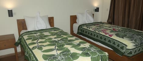 Quarto de hotel com duas camas de solteiro, cada uma com cabeceira de madeira, almofadas brancas e colcha estampada com flores verdes. Há duas mesas de cabeceira fixadas na parede, com acabamento em madeira, e uma cortina marrom na janela.
