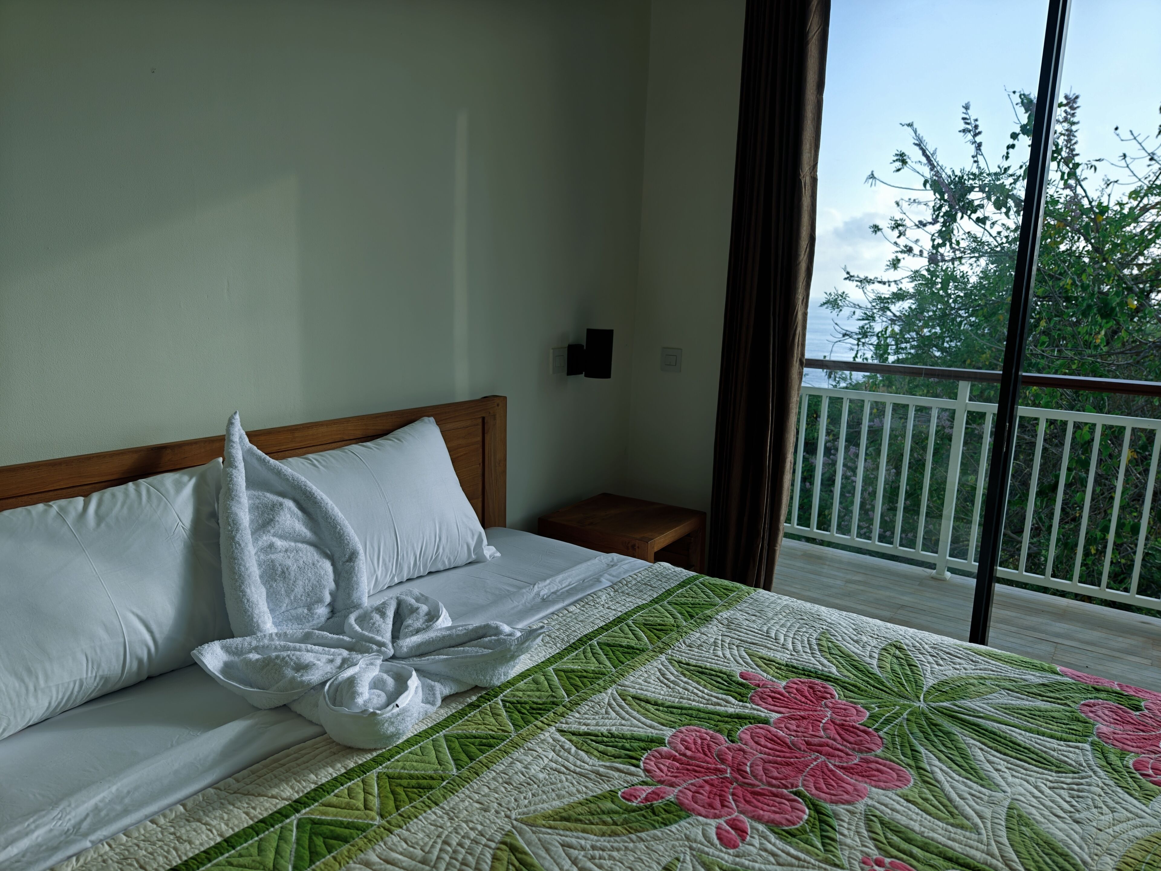 A hotel room with a bed, a nightstand, and a view of a balcony with a railing.