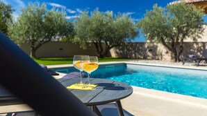 A poolside view with a table and a glass of drink, surrounded by trees and a clear sky.