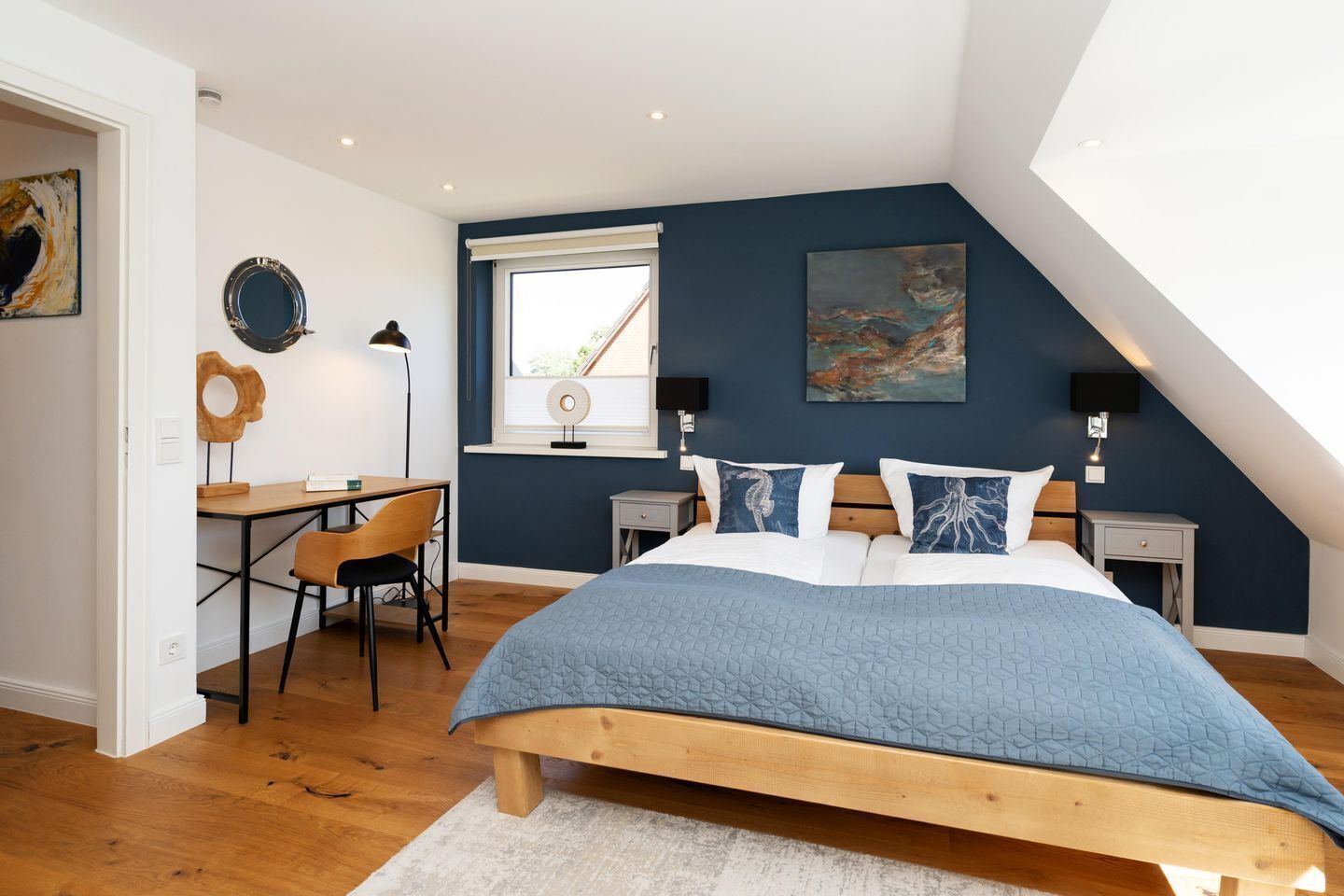 A bedroom with a sloped ceiling, a bed with a blue quilt, a desk with a chair, and a window with blinds.