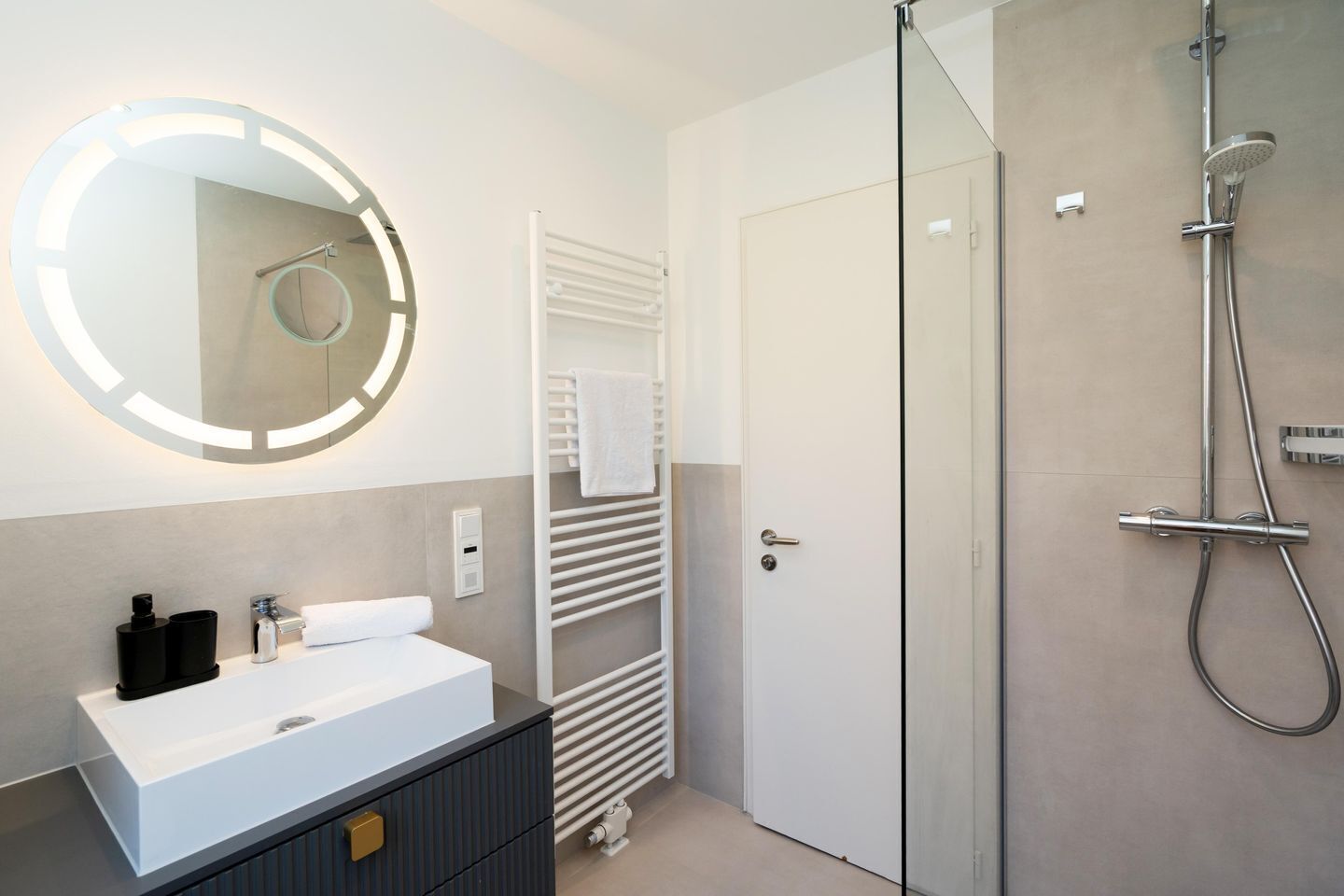 Ein modernes Badezimmer mit rundem Spiegel, Waschbecken in Weiß, Handtuchhalter, Dusche und einer Tür.