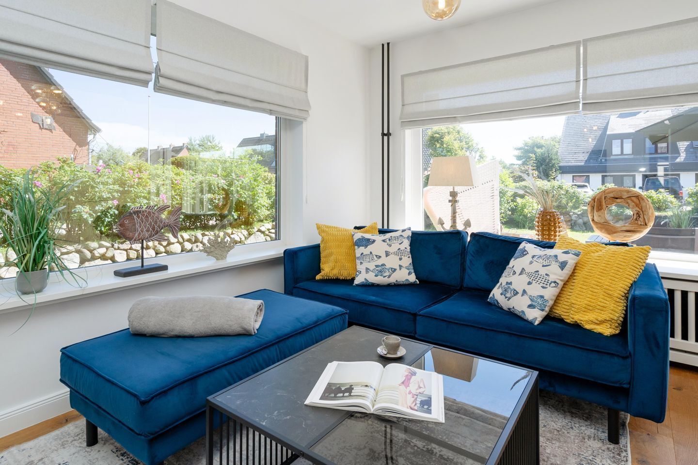 Een moderne woonkamer met een blauwe modulaire bank, een glazen salontafel en uitzicht op een tuin door grote ramen.