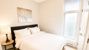 A bedroom with a bed, a bedside table with a lamp, a wall-mounted artwork, and a door to the right.