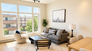 A modern living room with a sofa, armchair, coffee table, dining table, and a large window offering a view of buildings and trees.