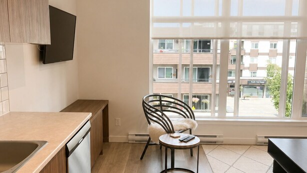 Une cuisine moderne avec des meubles en bois, un lave-vaisselle encastré, une table de salle à manger ronde avec une chaise, et une grande fenêtre donnant sur des immeubles.