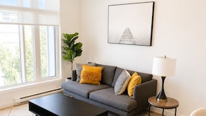 Un salon moderne comprenant un canapé, une table basse et un fauteuil, une table d’appoint avec une lampe, et une grande fenêtre donnant sur un paysage verdoyant.
