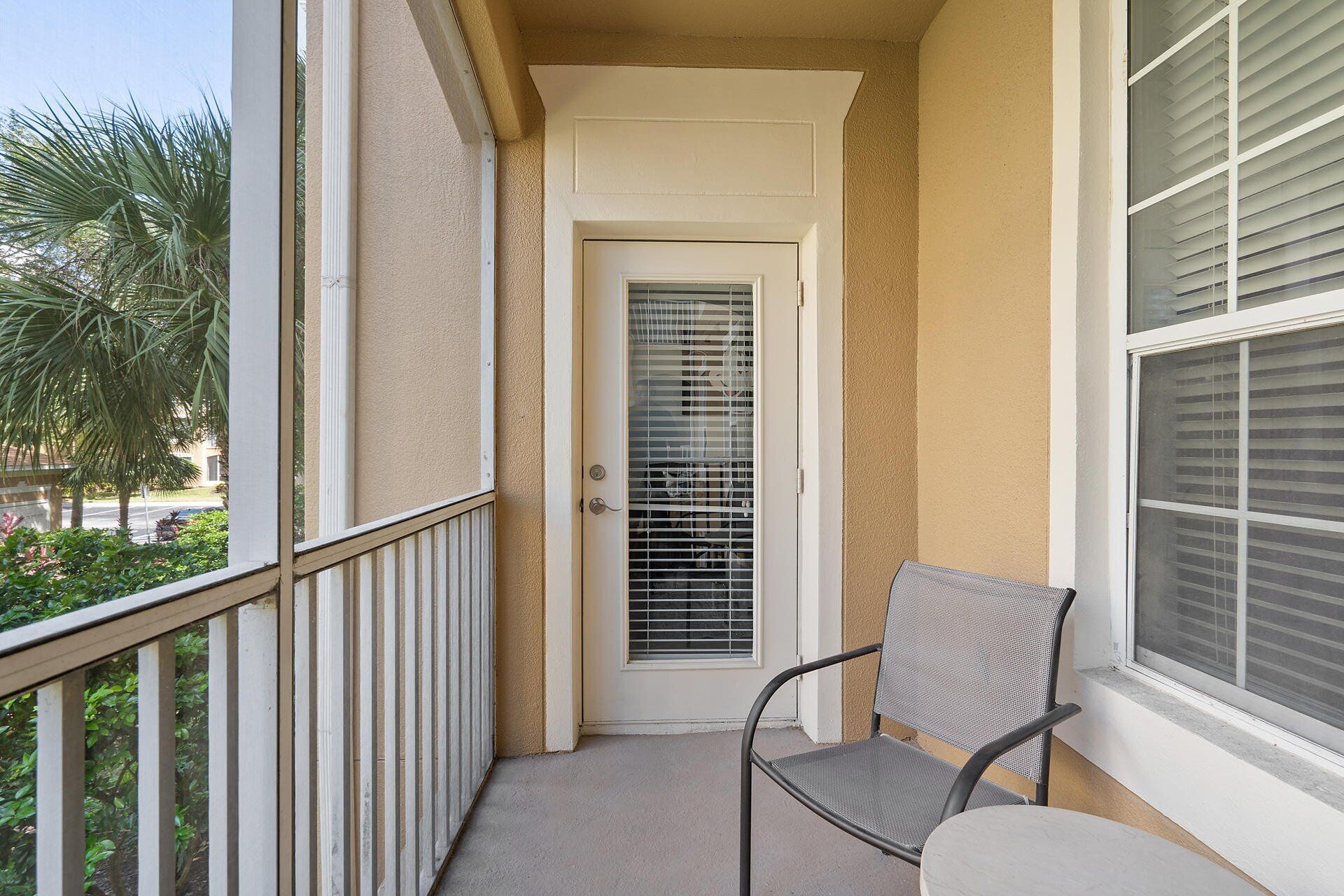 A balcony with a chair, a door with a window, and a view of palm trees.