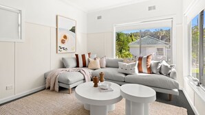 A modern living room with a grey sectional sofa, two white coffee tables, and a large window offering a view of a house and greenery.