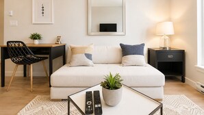 Una sala de estar moderna con un sofá, una mesa de centro con controles remotos, una mesita de noche con una lámpara, una silla, un escritorio y una planta en maceta.