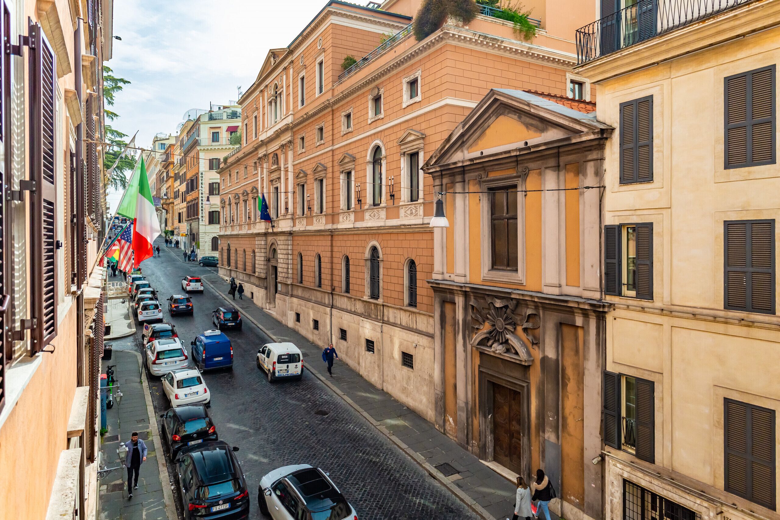 Una calle estrecha flanqueada por vehículos estacionados y edificios históricos con balcones y contraventanas.
